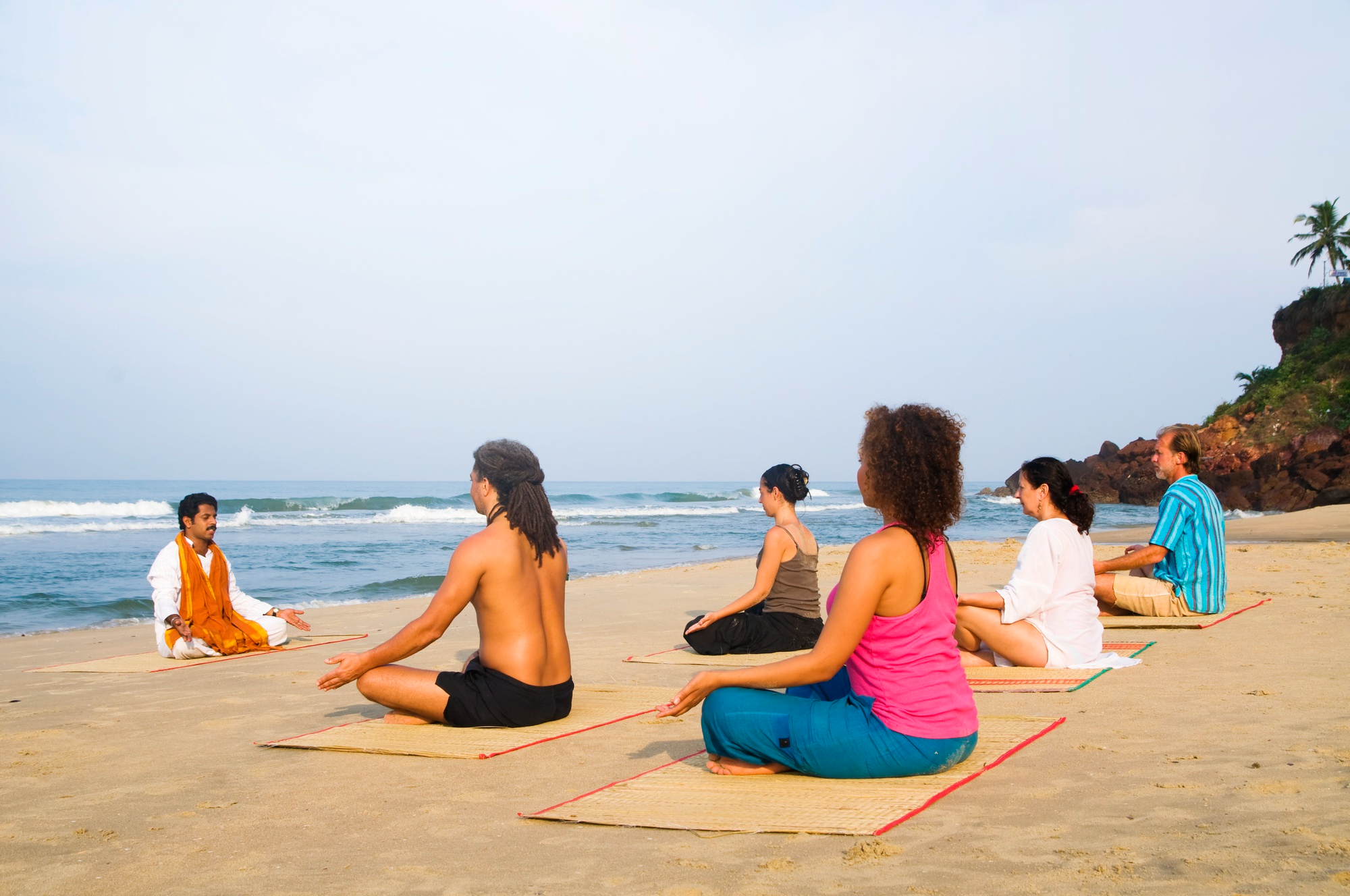 A group of people practising meditation on a beach at sunrise, sitting in a row on yoga mats facing the ocean.