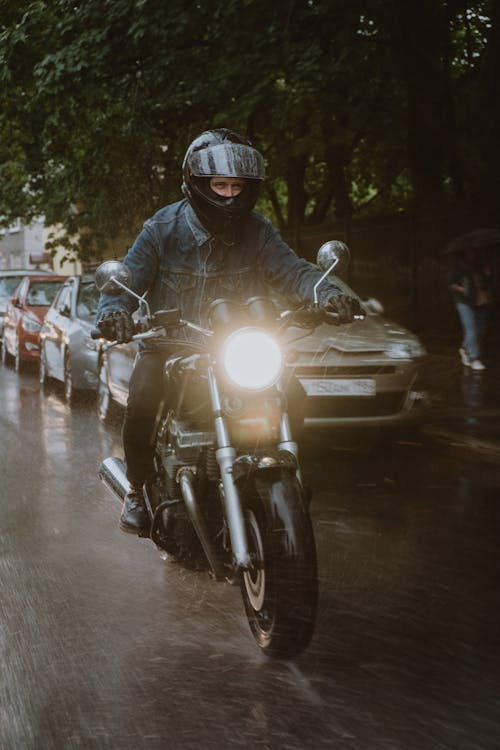 A person wearing a helmet and a dark jacket rides a motorcycle with its headlight on along a wet street during rainfall.