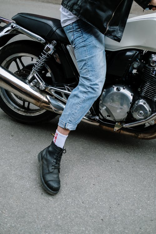 A person wearing light blue jeans, black boots, and a black leather jacket, sitting on a silver and black motorcycle