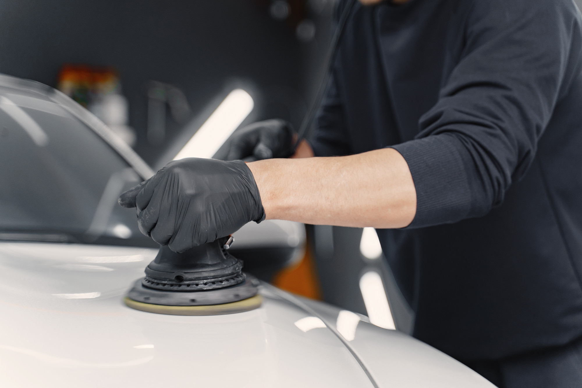 Close-up of a professional detailer wearing black gloves using a polishing machine on the hood of a white car in a garage.