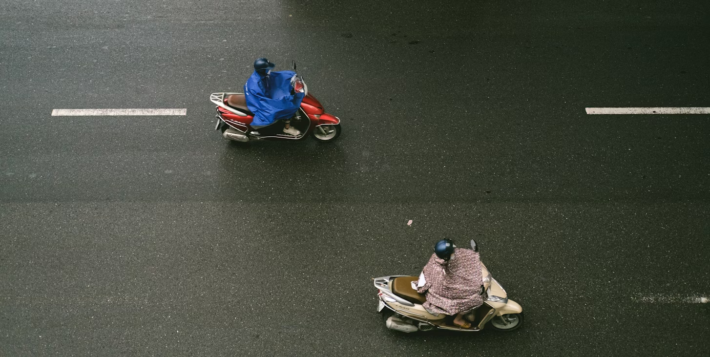Two riders on scooters, covered in raincoats, travel on a wet road.