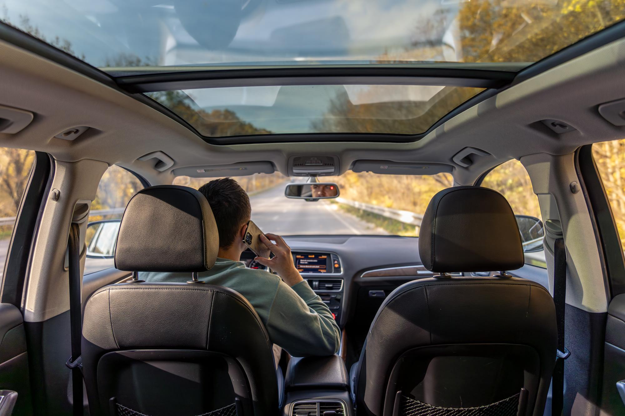 Interior view of a car driving on a sunny road, showing a large panoramic sunroof above the front and rear seats with the driver talking on the phone.
